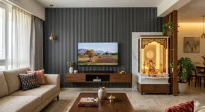 Contemporary Indian living room design featuring a statement TV unit wall with charcoal grey vertical fluted louvers, a floating wooden console, and a compact modern white marble pooja mandir with intricate CNC jaali woodwork and warm ambient backlighting.